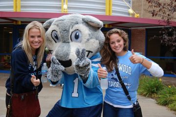 Two people in university shirts stand on either side of a costumed mascot giving thumbs up outdoors.