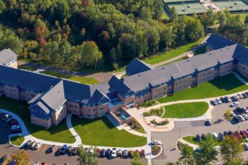 Aerial view of a large, L-shaped building surrounded by greenery. The building has a paved driveway and several parked cars, with tennis courts visible in the top right corner.