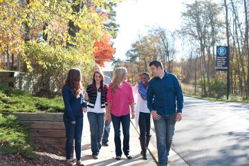 A group of six people, casually dressed, walking on a sidewalk through a park-like area on a sunny day. Trees with autumn foliage are in the background.