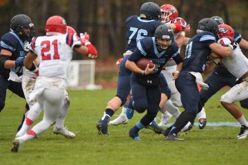 A football player in dark blue uniform runs with the ball while being pursued by opponents in white and red uniforms on a grassy field. Other players from both teams are engaged in blocking, showing the intense competition you can expect at any Point After Club event.