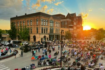 A large crowd gathers for an outdoor concert in Midland's charming town square at sunset, with a band performing on stage and historic brick buildings framing the scene. Discover Midland's vibrant culture as music fills the air.