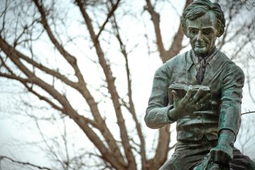 Bronze statue of a man reading a book, set against a backdrop of leafless tree branches.
