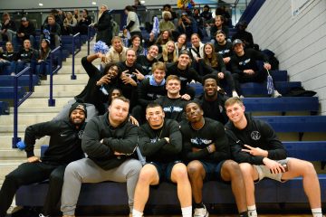 A group of people in casual sports attire sits on bleachers in a gymnasium, smiling and posing for the camera.