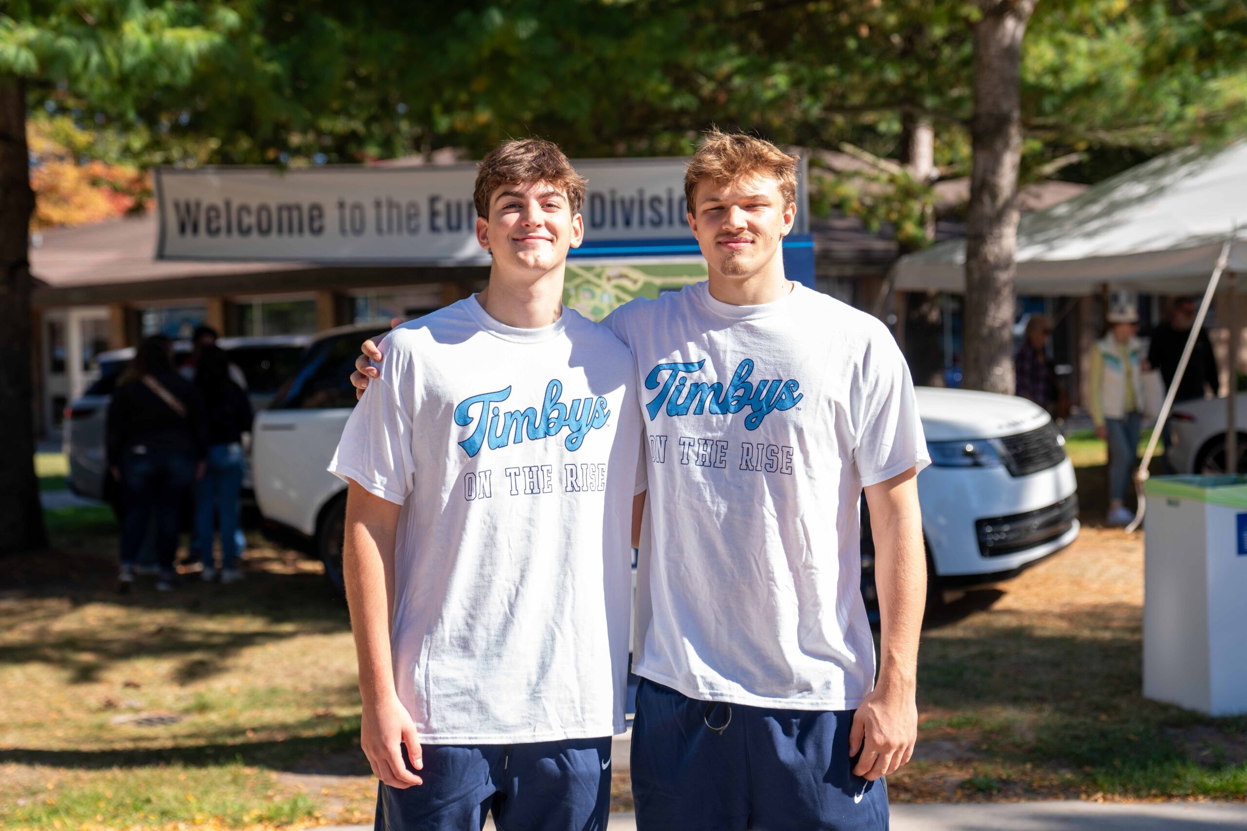 Two people stand side by side outdoors, wearing matching "Timber on the Rise" T-shirts. A "Welcome to the Europe Division" banner hints at a global outreach, bridging gaps between automotive education and innovation among nature's embrace. Trees in the background add to this cross-continental unity.